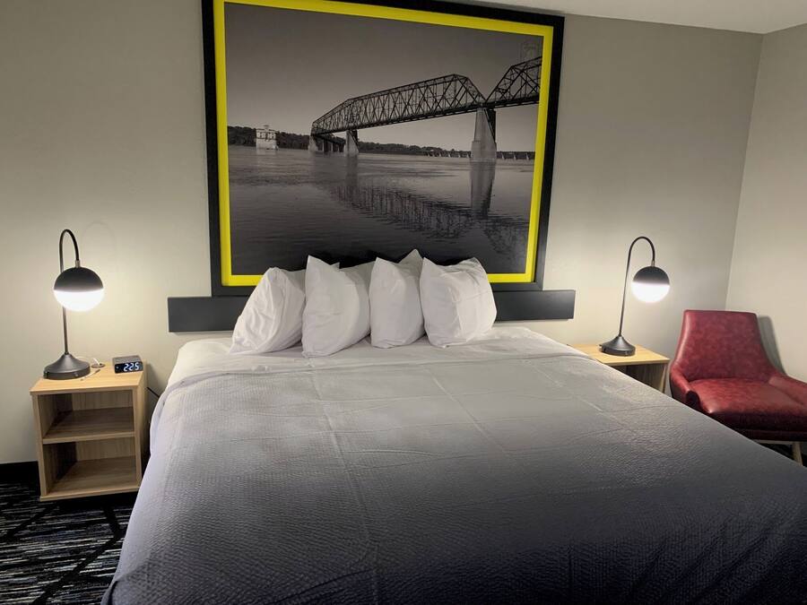 A hotel room with a large bed, two bedside lamps, a red chair, and a framed picture of a bridge.