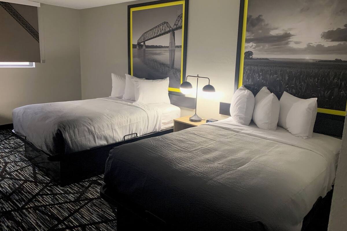 A hotel room with two beds, a nightstand with a lamp, and a window with blinds.