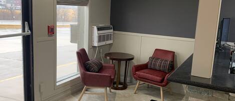 Una habitación pequeña y moderna con dos sillas rojas, una mesita redonda, un detector de incendios y una ventana con vistas al exterior.