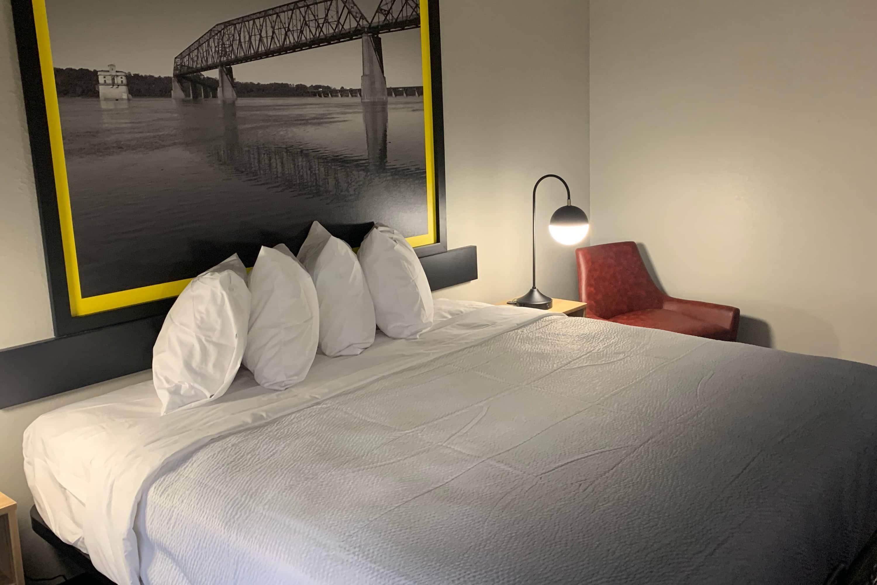 Quarto de hotel com cama, mesa de cabeceira, abajur, cadeira vermelha e grande quadro com imagem de uma ponte.