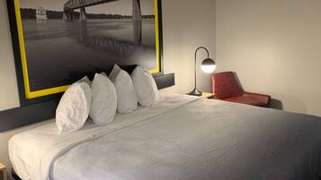 Quarto de hotel com cama, mesa de cabeceira, abajur, cadeira vermelha e grande quadro com imagem de uma ponte.
