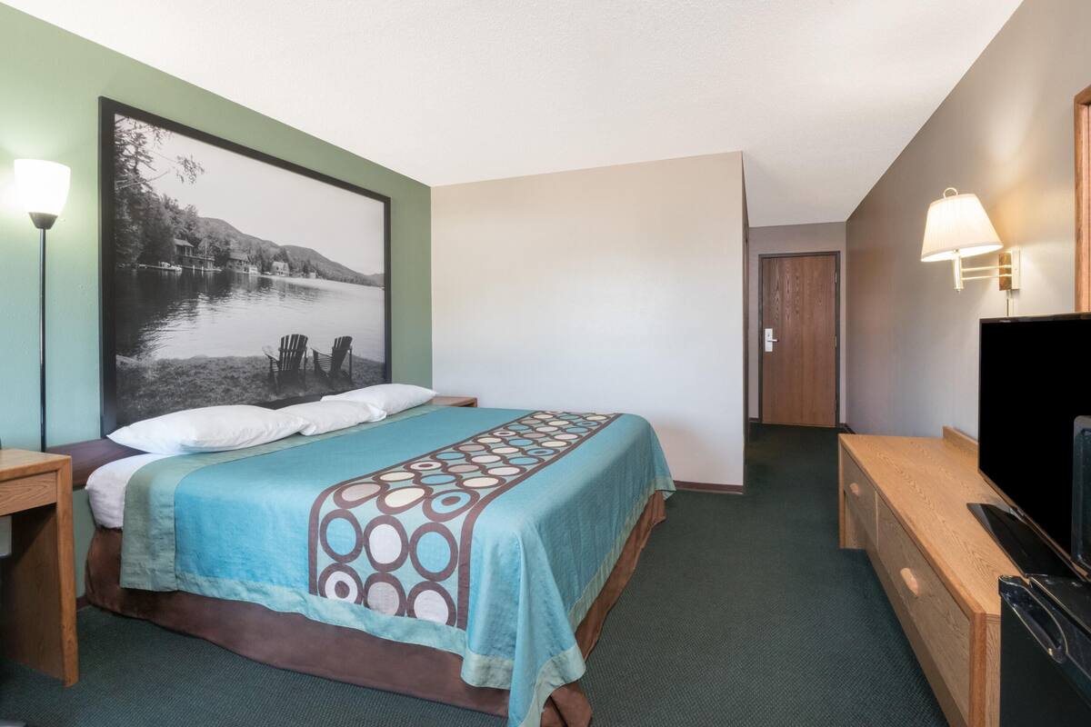A hotel room with a bed, a TV, a desk, and a large framed picture on the wall.