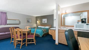 A compact hotel room with a bed, a desk with a chair, a small kitchenette, a sofa, and a bathroom.