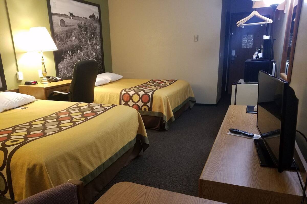 A hotel room with two beds, a desk, a chair, a TV, and a lamp.