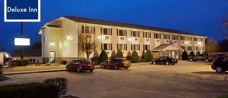 Un hotel con un cartel que dice "Deluxe Inn", que cuenta con un estacionamiento con varios coches, un edificio con múltiples ventanas y una entrada bien iluminada.