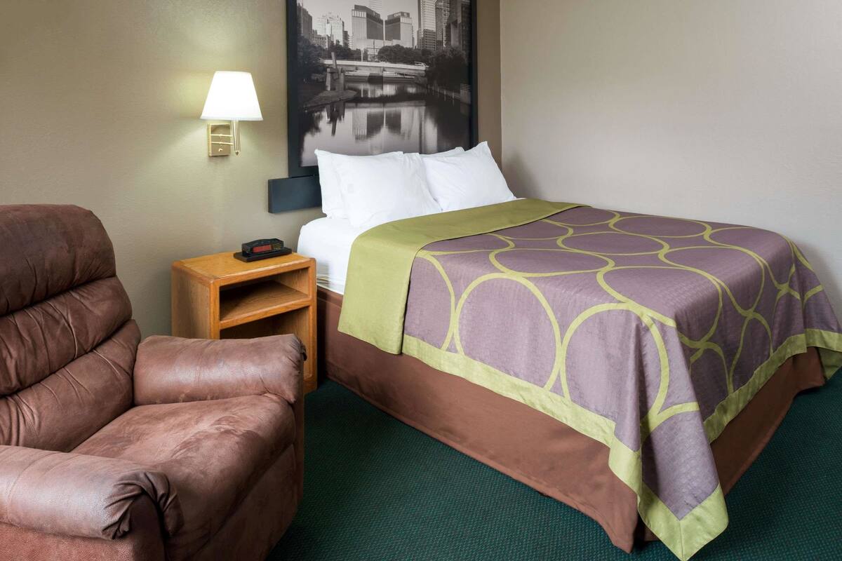 Une chambre d’hôtel avec un lit, un fauteuil en cuir marron, une table de chevet avec une lampe et un tableau représentant un paysage urbain accroché au mur.