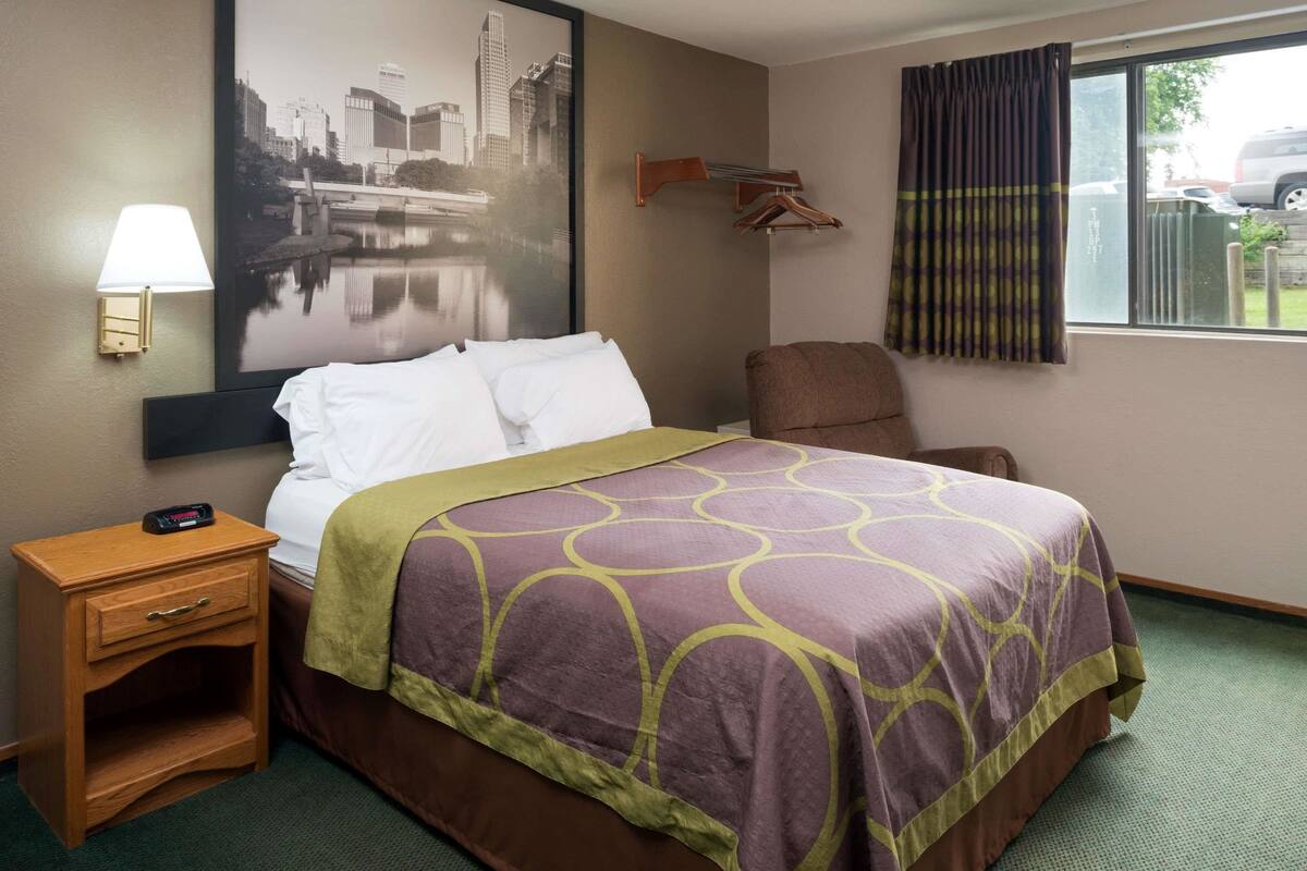 Une chambre d’hôtel comprenant un lit, une table de chevet, une chaise, une lampe fixée au mur, un porte-manteau et une fenêtre avec des rideaux.