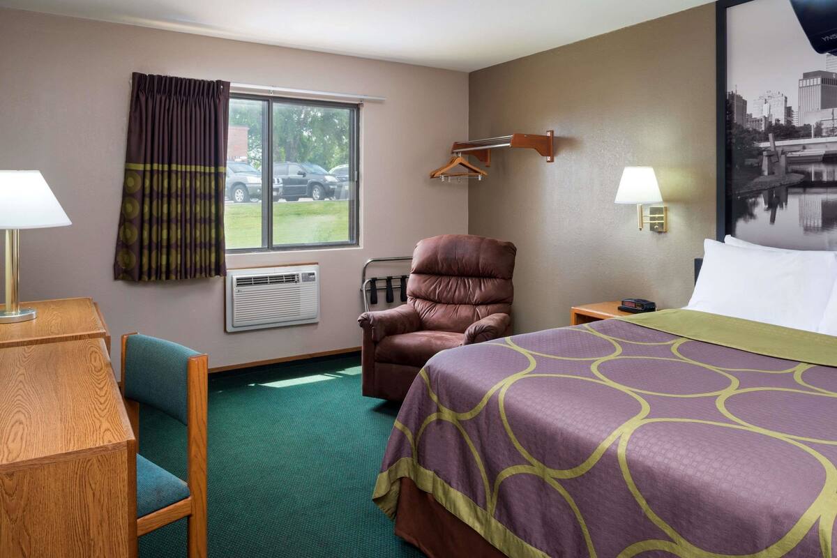 Une chambre d’hôtel comprenant un lit, une chaise, un bureau, une fenêtre avec des rideaux, un appareil de climatisation, une lampe fixée au mur et un porte-manteau.