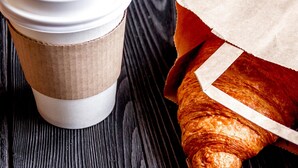 A coffee cup with a lid and a paper bag containing a croissant on a wooden surface. - Americas Best Value Inn Seward (Seward)