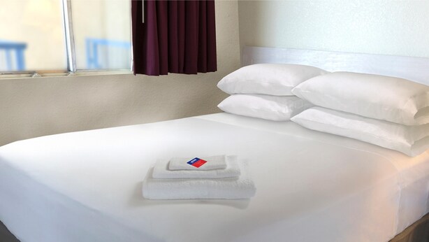 A neatly made bed with white linens and pillows, a folded towel, and a window with curtains. - Americas Best Value Inn Seward (Seward)