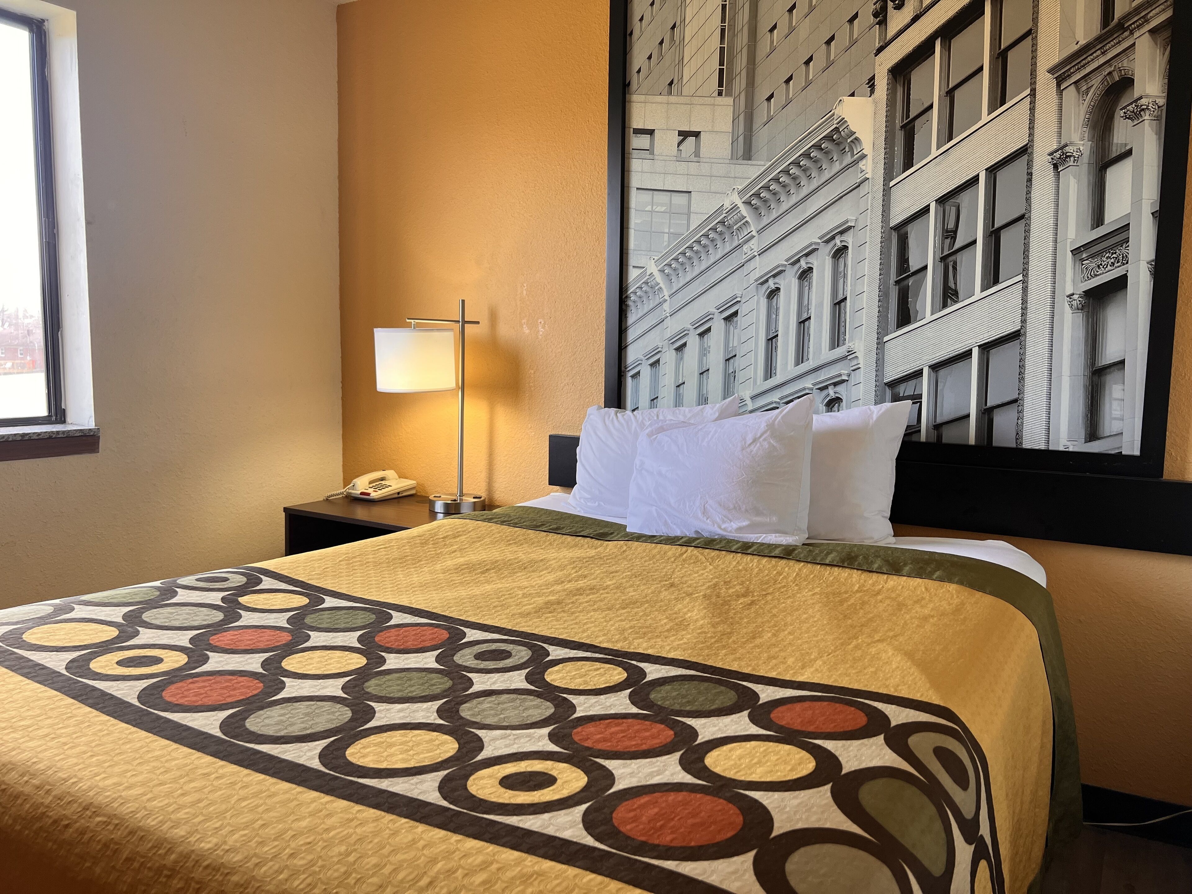 Een hotelkamer met een bed, nachtkastje, lamp en een groot raam met uitzicht op de stad.