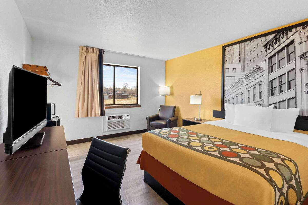 A hotel room with a bed, a desk with a computer, a chair, a window with curtains, and a lamp.