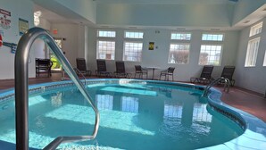 Une piscine intérieure avec un escalier en métal, entourée de chaises et de tables, et de grandes fenêtres laissant entrer la lumière naturelle.