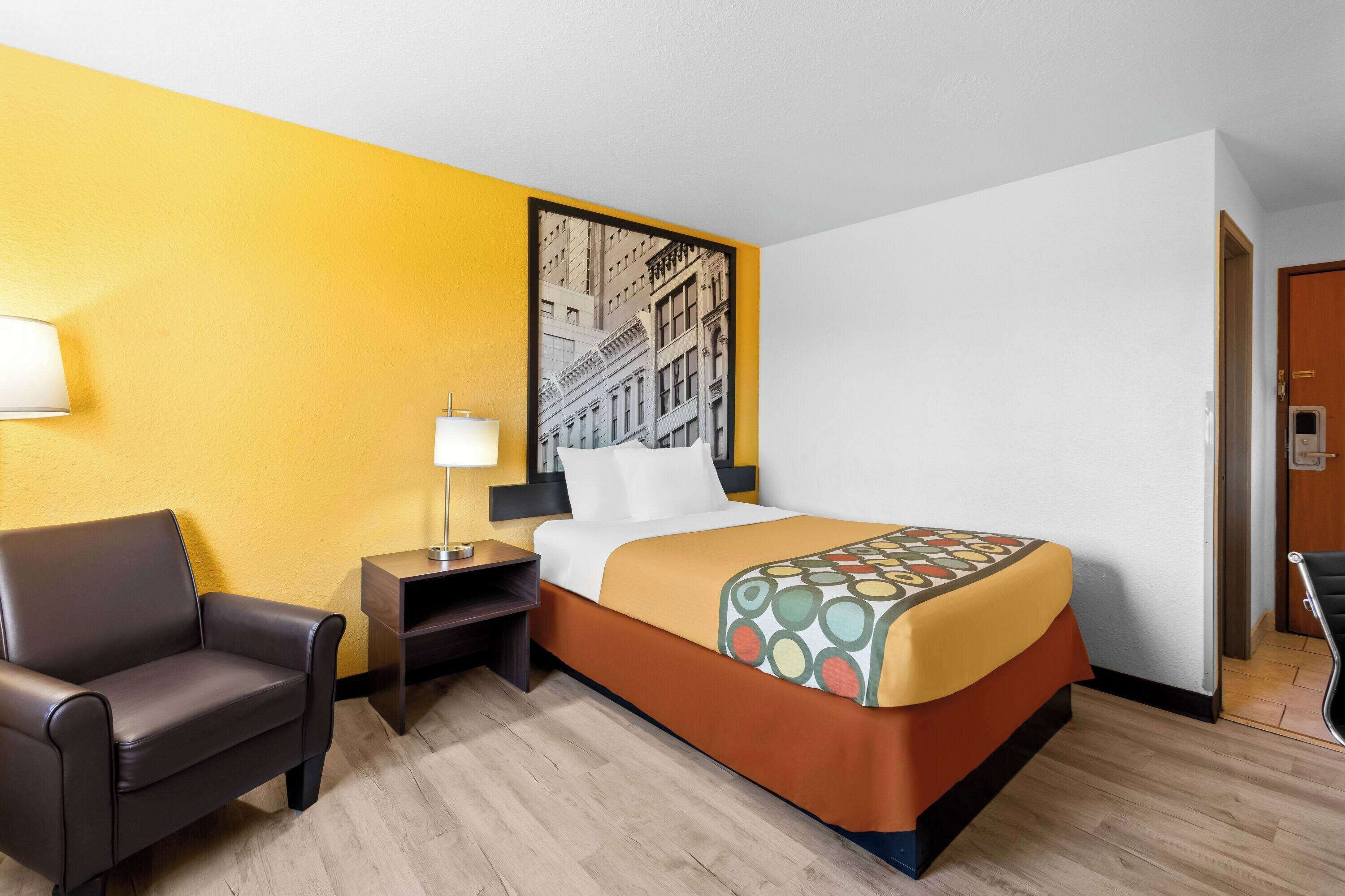 Une chambre d’hôtel avec un lit, une chaise, une table de chevet et un mur orné d’un tableau représentant un paysage urbain.