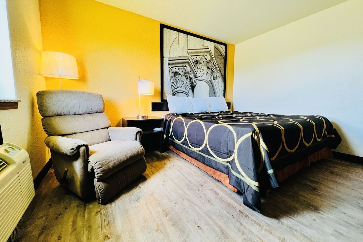 A hotel room with a bed, a chair, a nightstand, a lamp, and a radiator.