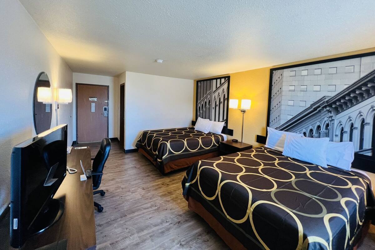 A hotel room with two beds, a desk, a TV, and a wall-mounted lamp.