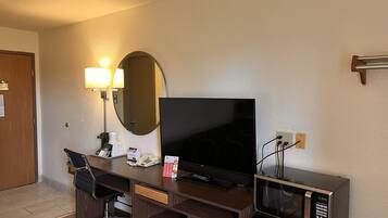 A hotel room with a desk, a flat-screen TV, a microwave, a desk lamp, and a mirror.
