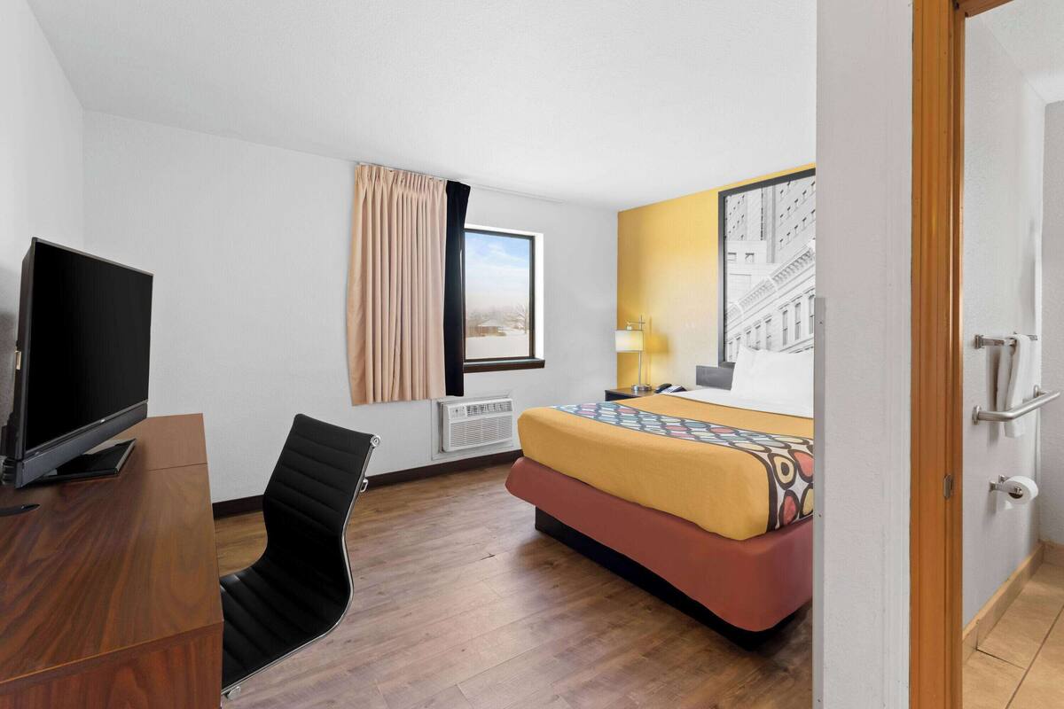 A hotel room with a bed, a desk with a TV, a chair, a window with curtains, and a wall-mounted lamp.