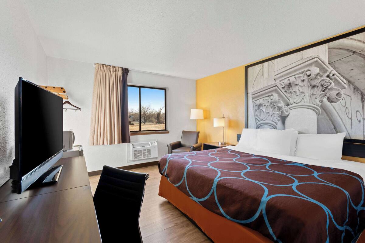 A hotel room with a bed, a desk with a flat-screen TV, a chair, a window with curtains, a lamp, and a wall-mounted air conditioner.