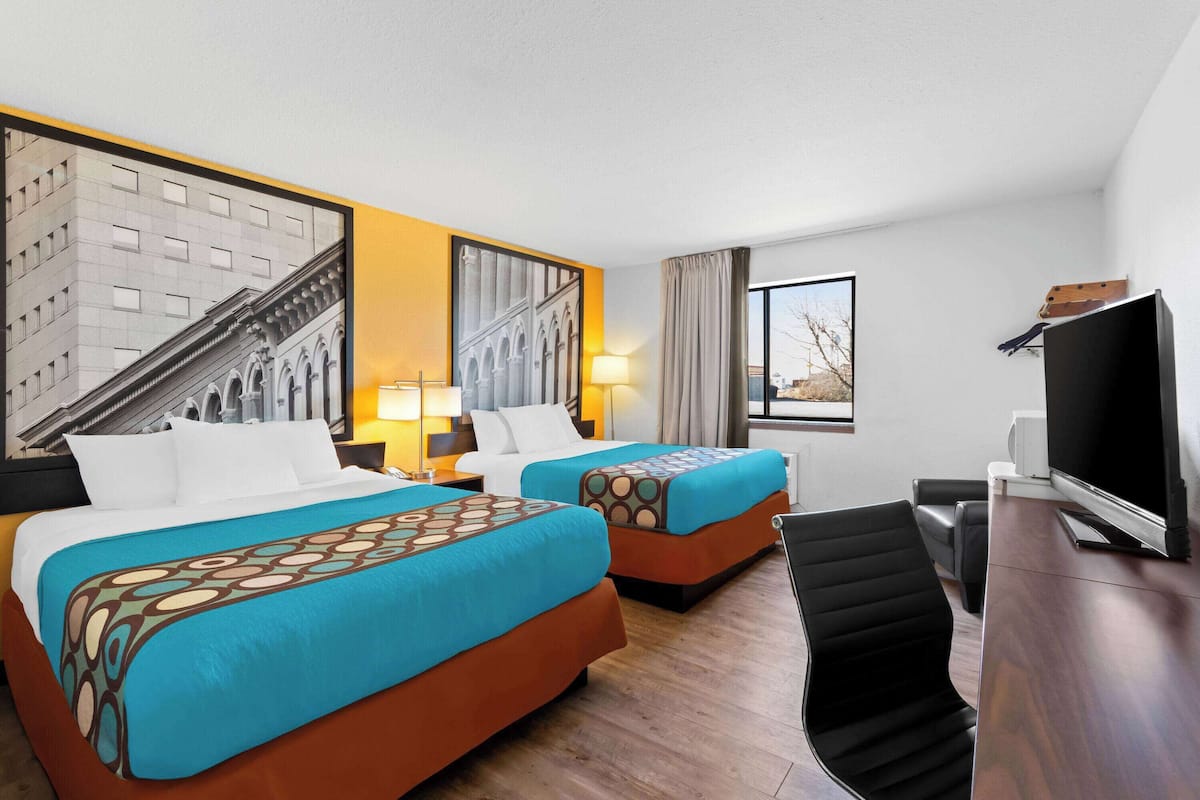 A hotel room with two beds, a desk, a TV, and a large window with a view of a building.