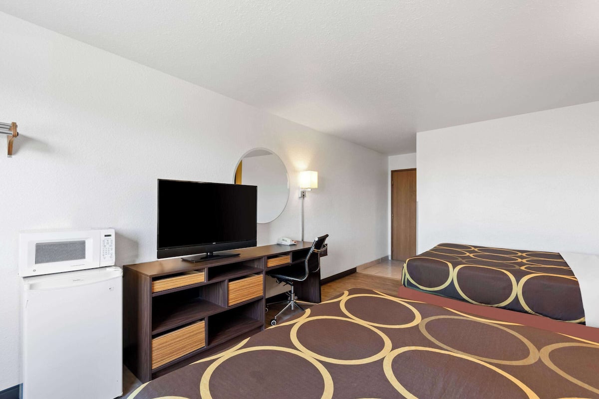 A hotel room with a flat-screen TV, a microwave, a desk with a chair, and two beds with patterned bedding.