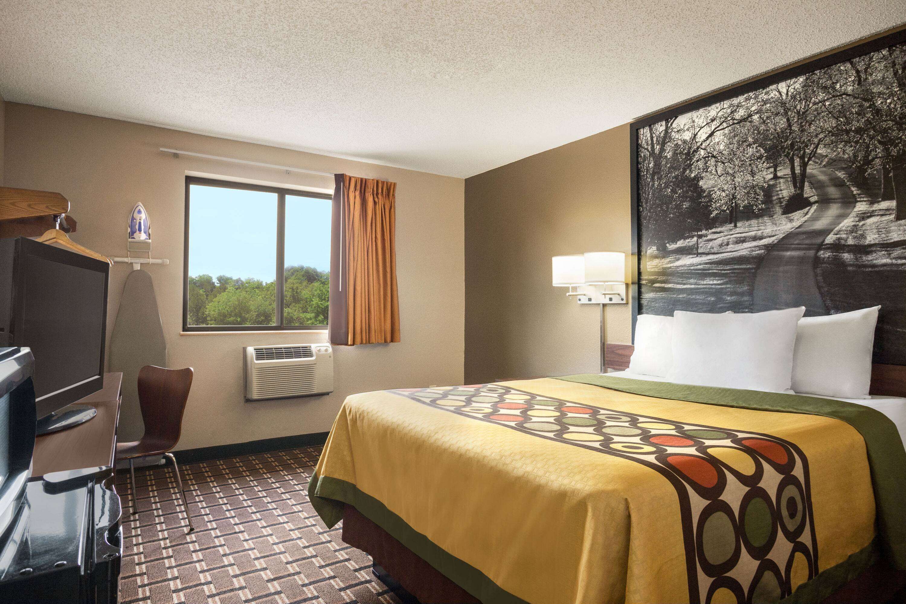 A hotel room with a bed, a desk with a television, a chair, a window with curtains, an air conditioner, and a wall-mounted lamp.