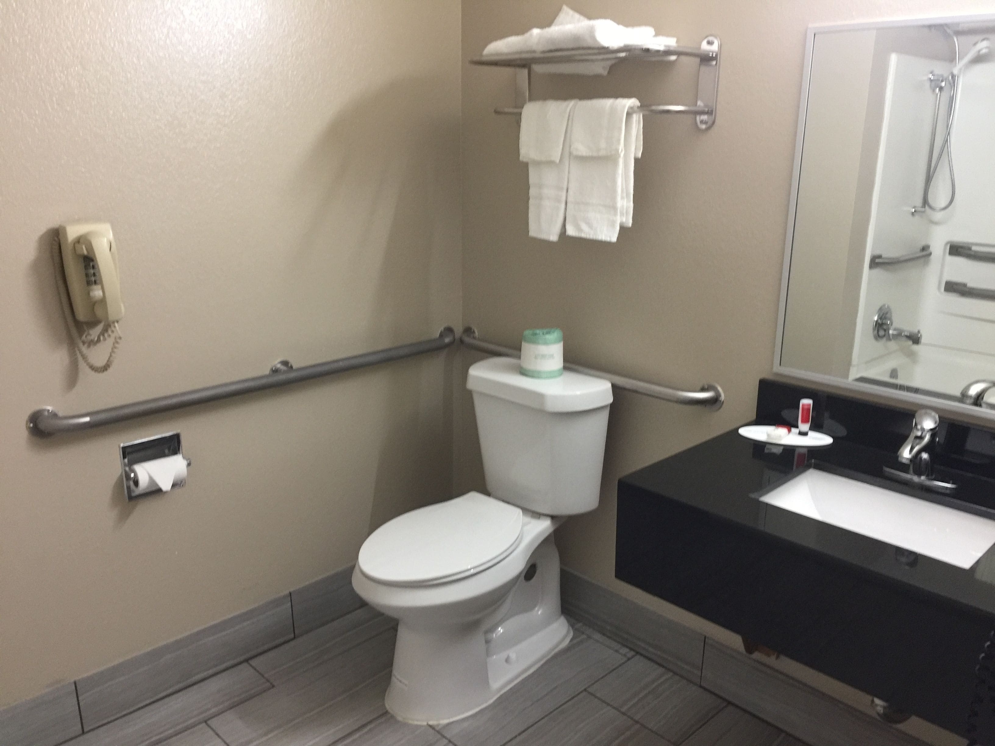A bathroom with a white toilet, a sink with a black countertop, a towel rack with towels, a handrail, and a mirror.