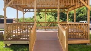 Un belvédère en bois doté d’une terrasse couverte et de côtés ouverts, entouré de verdure.