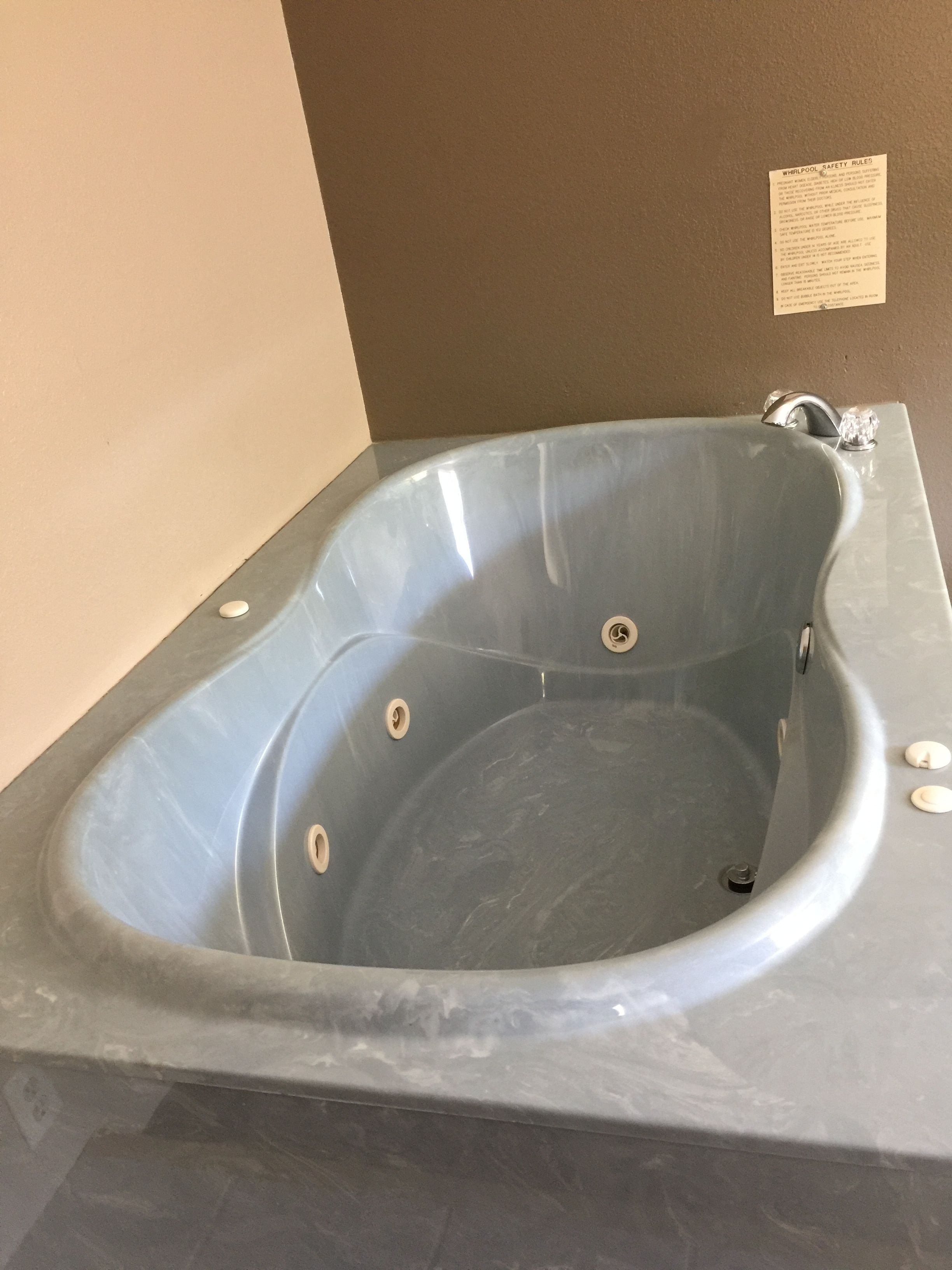Una bañera grande y rectangular, con acabado de mármol y borde curvo, ubicada frente a una pared color beige con un pequeño letrero de seguridad.