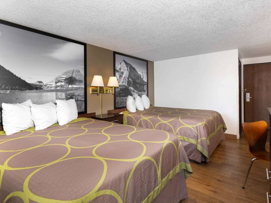 Ein Hotelzimmer mit zwei Betten, einem Schreibtisch und einem Sessel. Zwei Wandlampen und ein großes Bild einer Berglandschaft sind vorhanden.