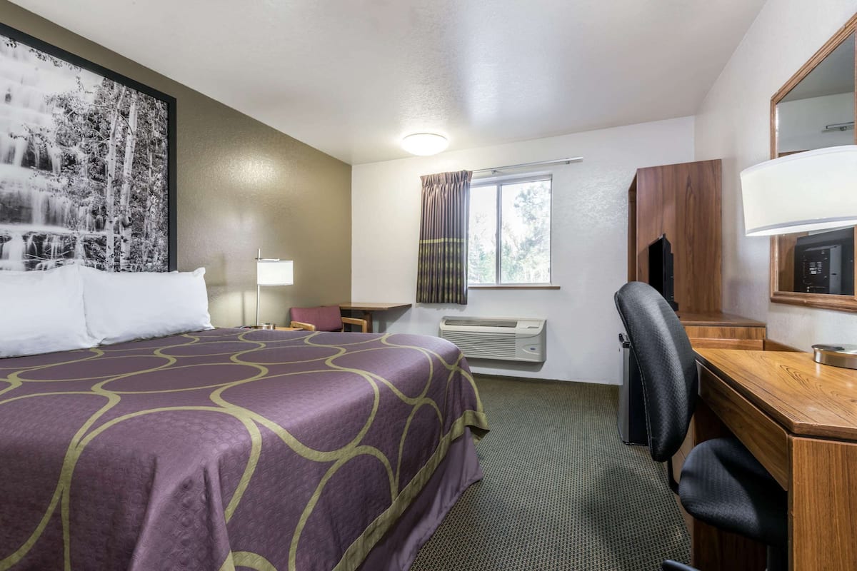 A hotel room with a large bed, a desk, a chair, a desk lamp, a window with curtains, an air conditioning unit, and a wall-mounted TV.