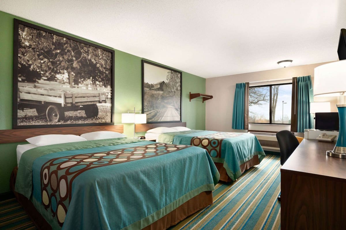 A hotel room with two beds, a desk, a chair, a lamp, and a large window with curtains.