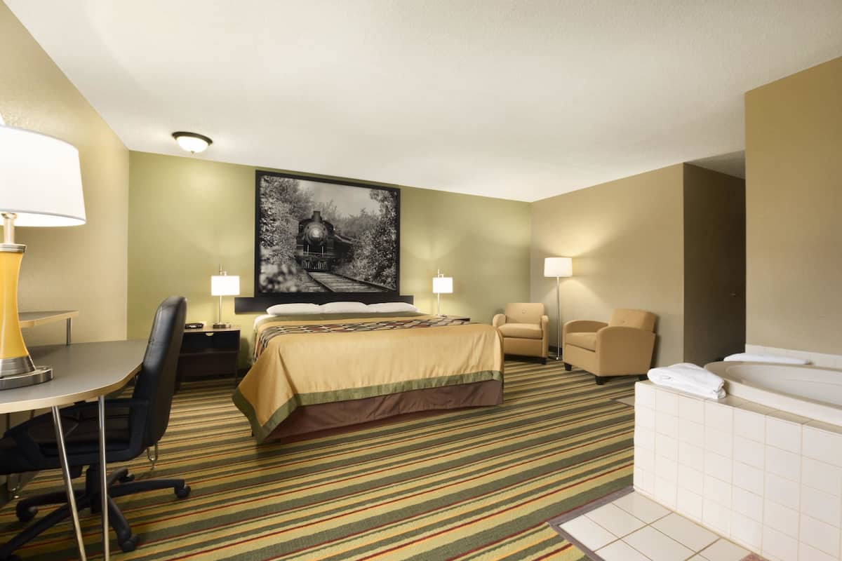 A hotel room with a bed, a desk, a chair, a desk lamp, a chair, a wall-mounted lamp, a bathtub, and a large painting on the wall.