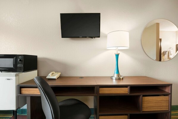 A hotel room with a desk, a flat-screen TV, a lamp, a microwave, a phone, and a mirror.