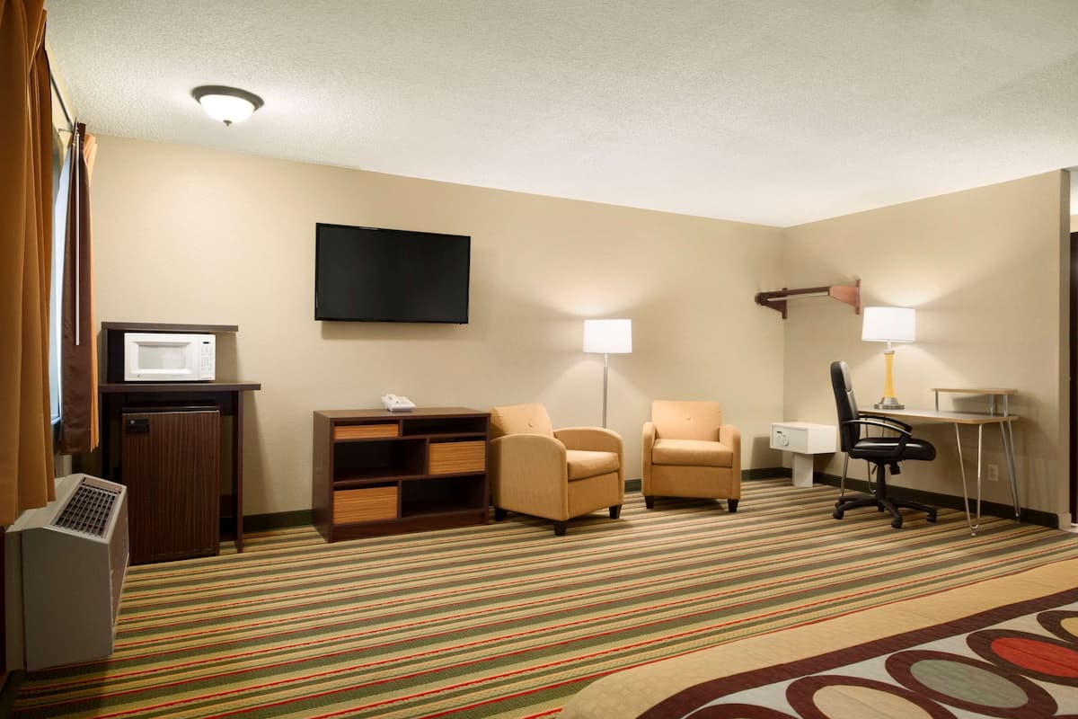A hotel room with a flat-screen TV, a microwave, a desk with a lamp, two armchairs, a small cabinet, and a striped carpet.