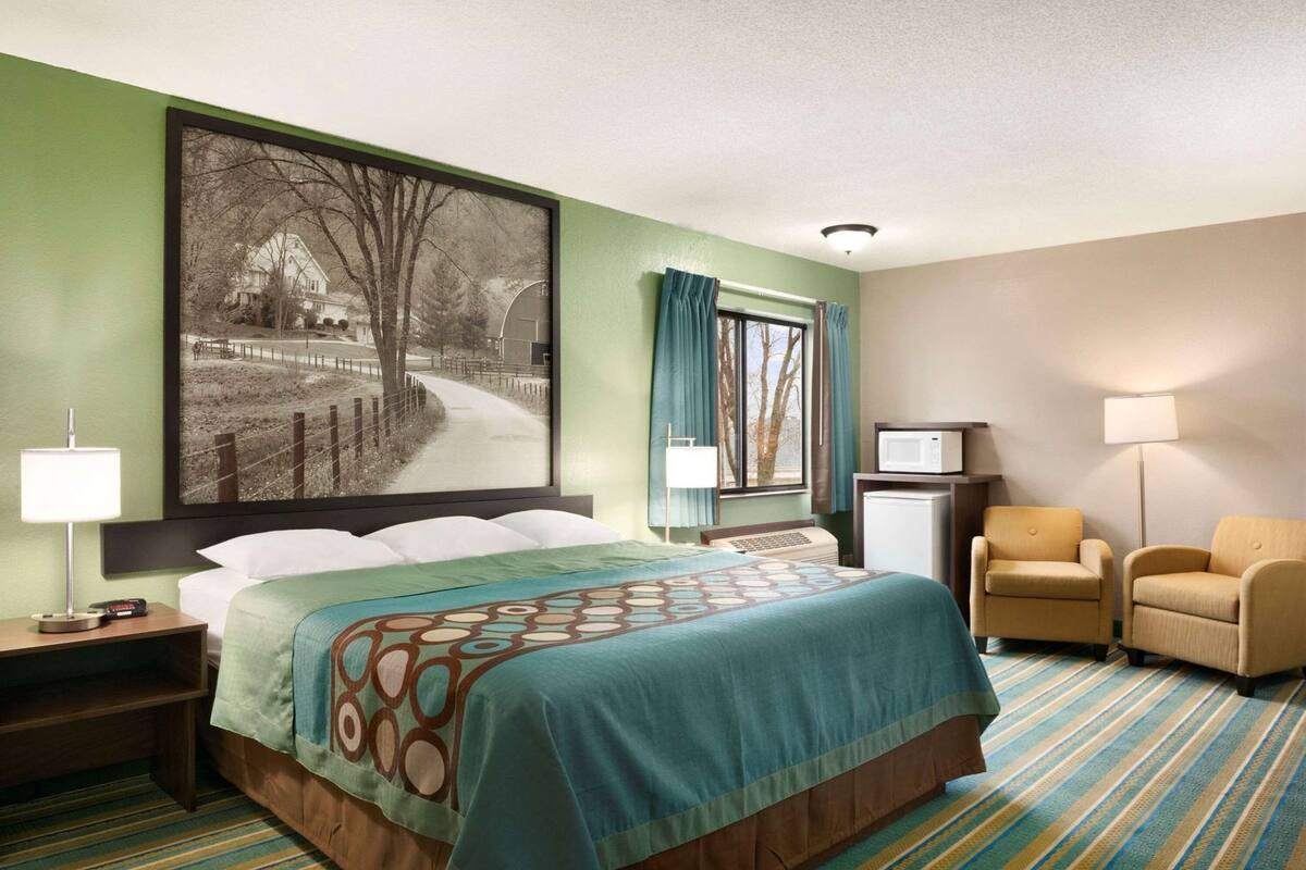 A hotel room with a bed, a desk with a lamp, a chair, a television, and a large window with curtains.