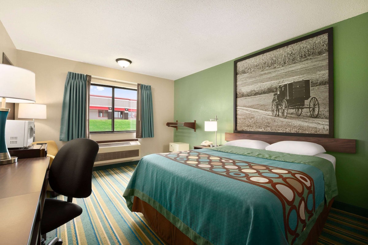 A hotel room with a bed, a desk, a chair, a window with curtains, a lamp, and a painting of a horse-drawn carriage.