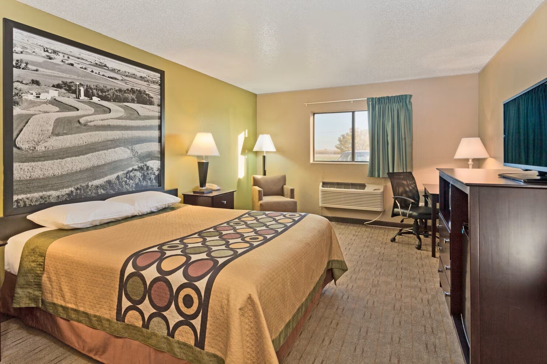 A hotel room with a bed, desk, chair, and a large landscape painting on the wall.