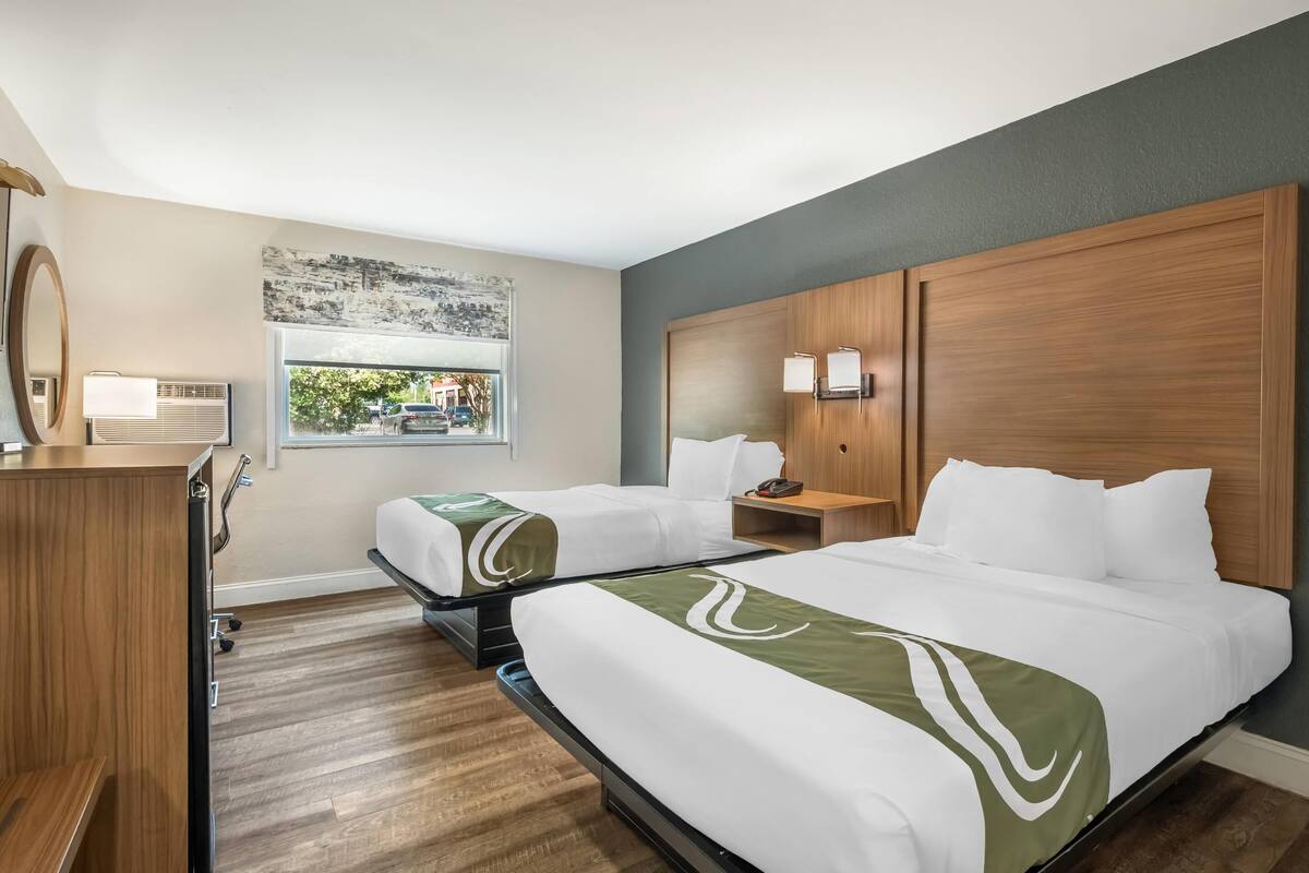 A hotel room with two beds, a desk, a chair, a window with a view of the outside, and a wooden headboard.