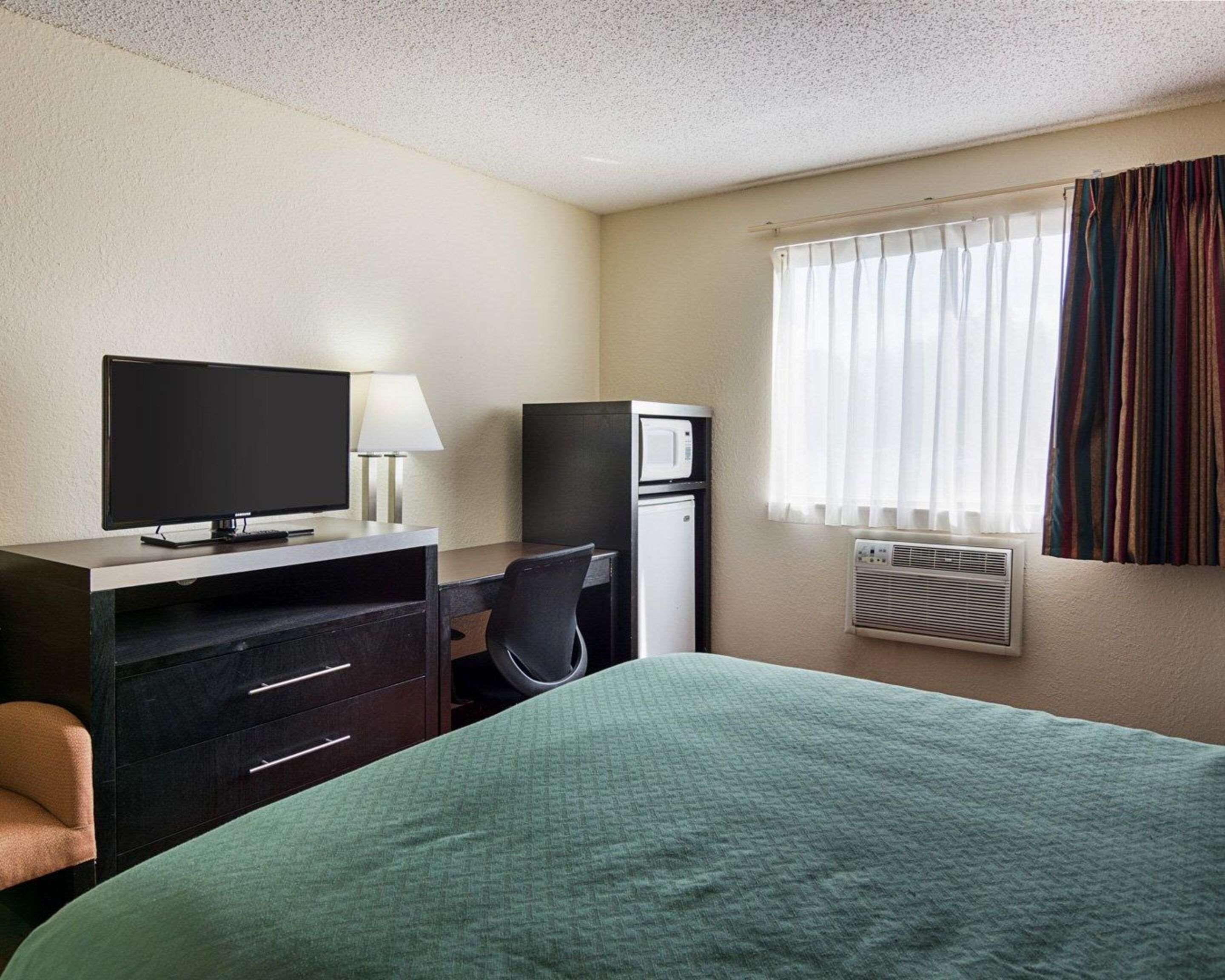 Een hotelkamer met een bed, een bureau met computer, een televisie, een koelkast en een raam met gordijnen.