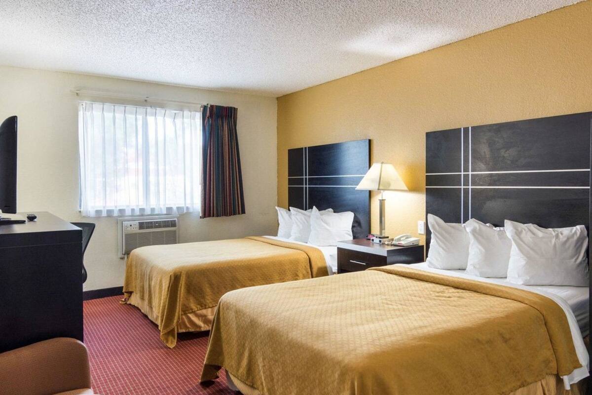 Quarto de hotel com duas camas, uma escrivaninha, televisão e janela com cortinas.