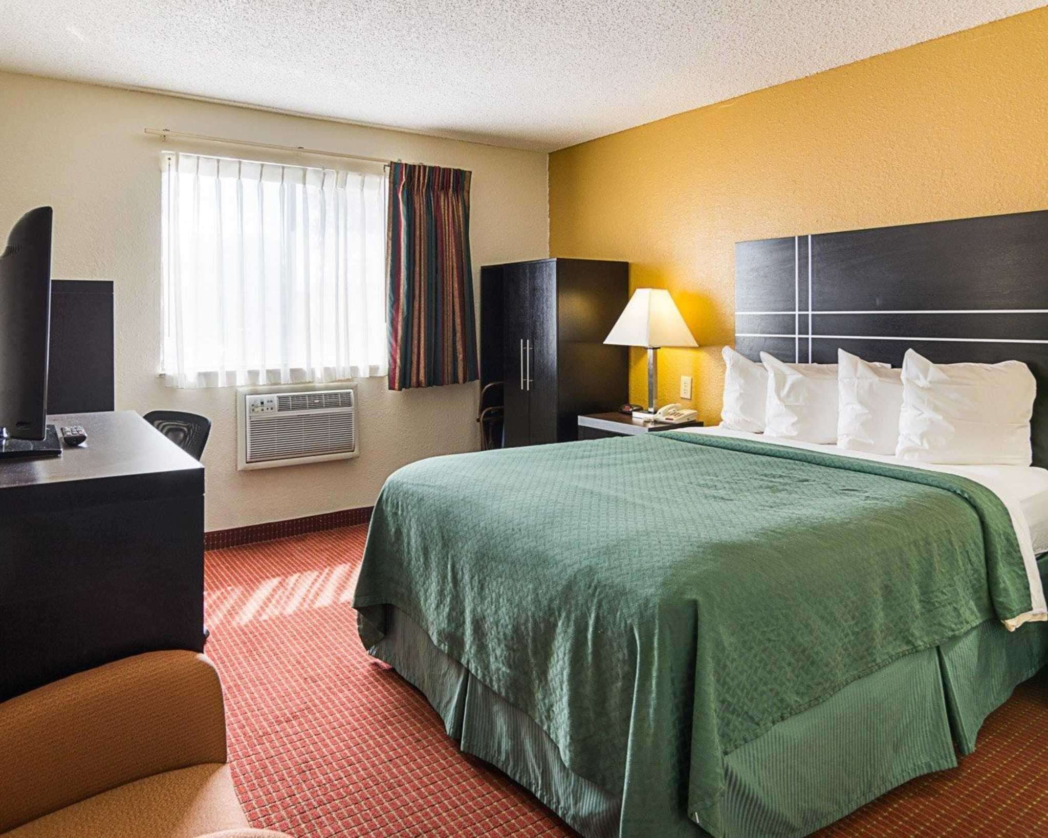A hotel room with a bed, a desk, a chair, a lamp, and a telephone.