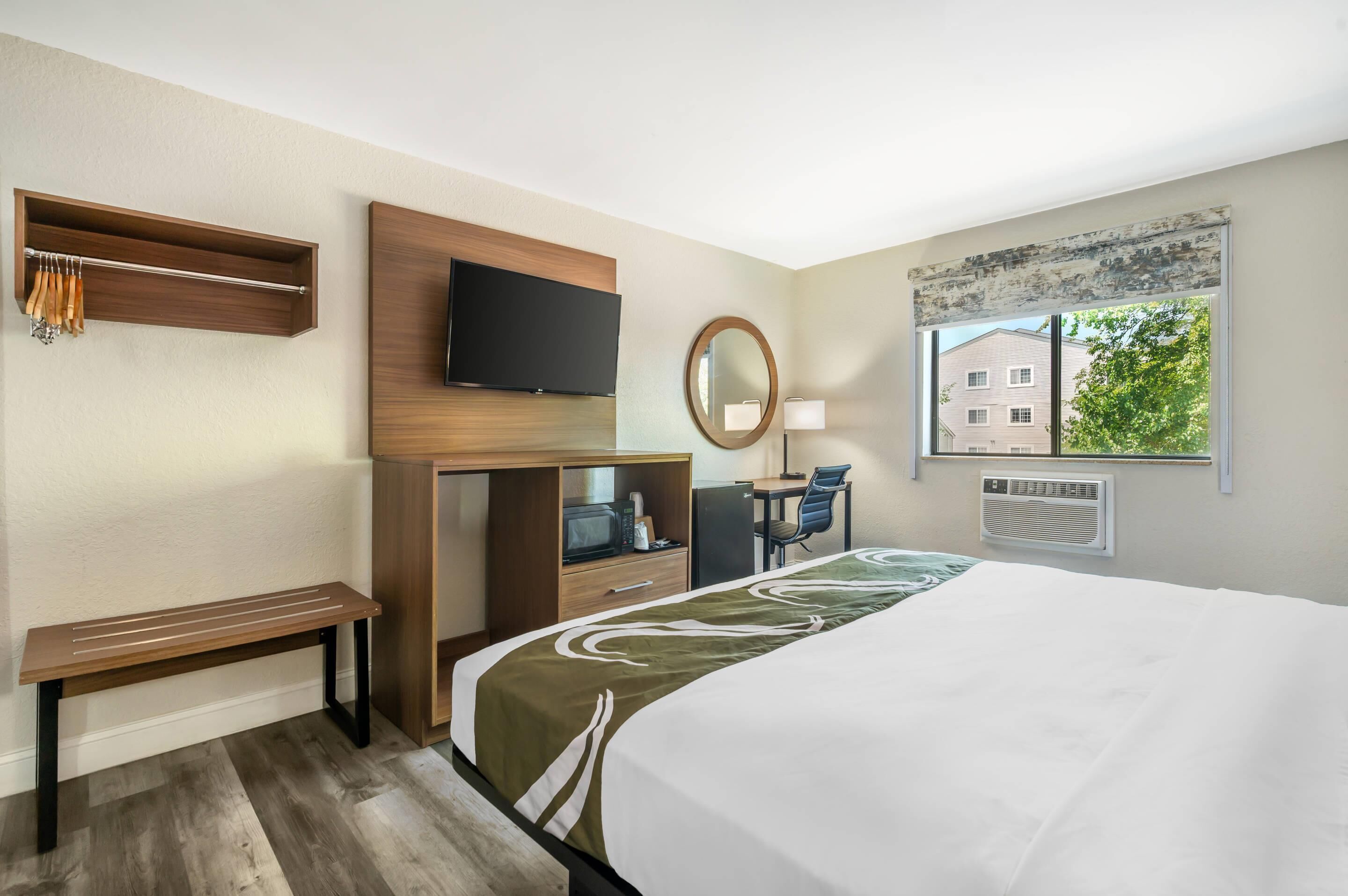 A hotel room with a bed, a desk with a chair, a television, a mirror, a window with a view of buildings, and a bench.