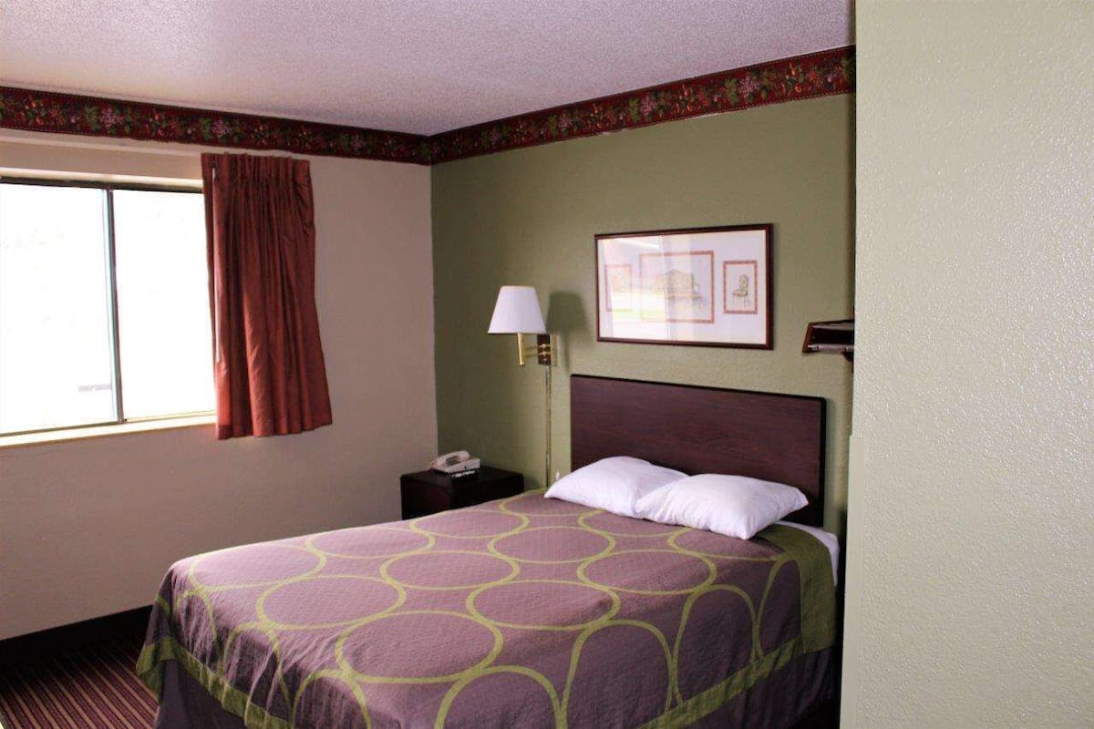 Quarto de hotel com uma cama, mesa de cabeceira, luminária, quadro na parede e janela com cortinas.