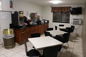 Cocina pequeña y limpia con refrigerador, cafetera y un basurero. Hay dos mesas y sillas, y una ventana con persianas.