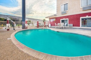 Una piscina con solárium azulejado, rodeada por una valla y sillas blancas.
