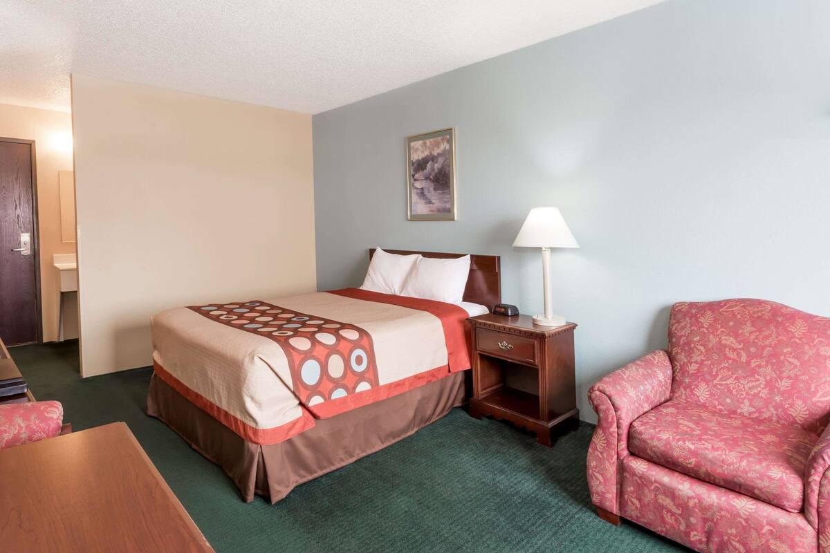 A hotel room with a bed, a nightstand, a chair, and a painting on the wall.
