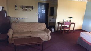 A living room with a sofa, a small table, a bed, a desk with a chair, a mirror, and a door.
