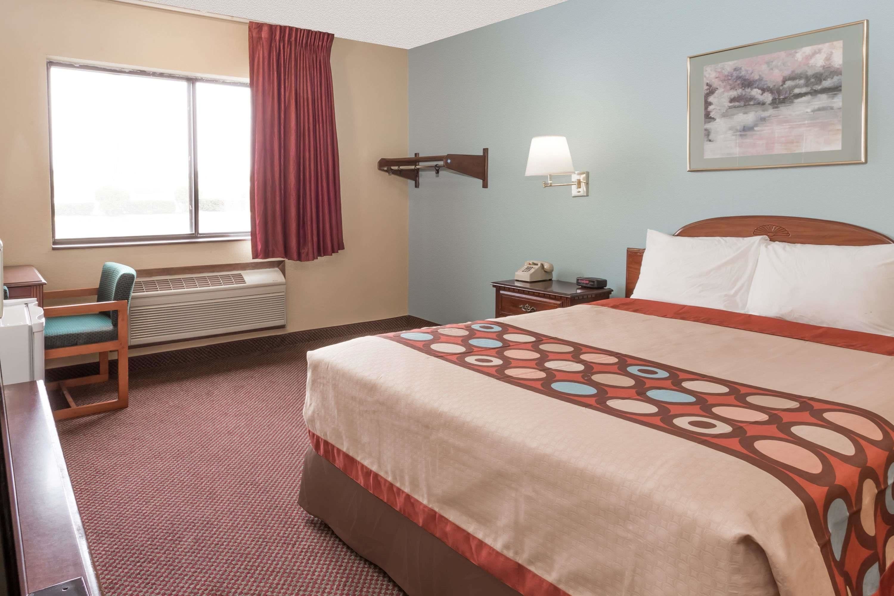 A hotel room with a bed, a desk, a chair, a window with curtains, a wall-mounted lamp, and a painting.