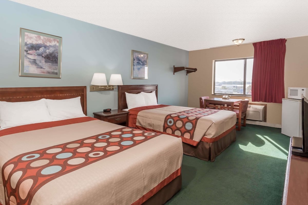 A hotel room with two beds, a desk, a chair, a TV, and artwork on the walls.